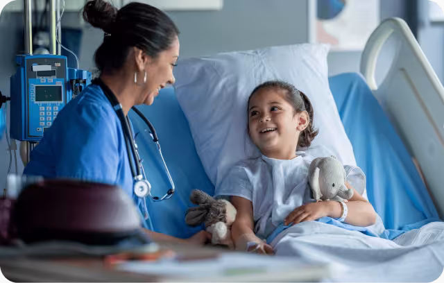 A nurse in blue scrubs smiling and talking with a young girl patient in a hospital bed holding stuffed animals