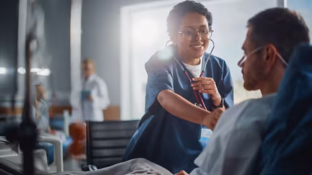 A smiling nurse in blue scrubs holding a stethoscope while attending to a male patient in a hospital bed