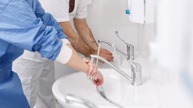 Two healthcare workers in scrubs washing their hands side by side at a clinical sink