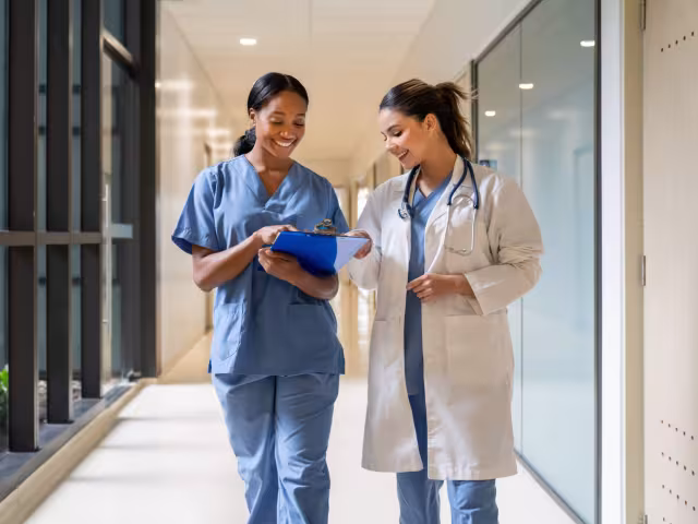 A nurse and doctor walking together down a hospital corridor while reviewing a clipboard